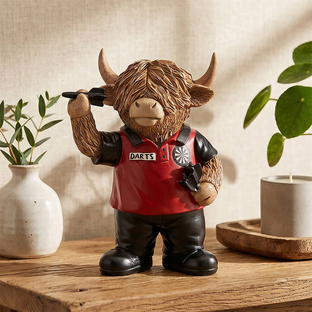 Grappig Schotse hooglander beeldje als darter met lange bruine vacht, rood-zwart dartshirt, zwarte broek en dartpijlen in zijn hoeven